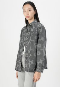 Chemise en denim grise avec un motif floral, fermeture à boutons et une poche poitrine unique ; coupe décontractée avec des manches longues et une texture douce.