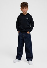 Zwarte hoodie met kangoeroezak en logo, gecombineerd met loszittende donkerblauwe jeans en witte sneakers met accentkleuren.