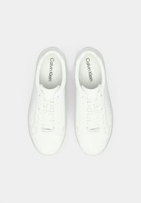 White sneakers with textured sides, lace-up design, and a smooth toe. Interior features brand name "Calvin Klein" in black.