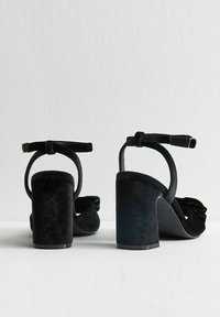 Zwarte fluwelen haksandalen hebben een dikke blokhak, open teenontwerp, enkelband met gespsluiting en een gerimpeld detail op de voorkant.