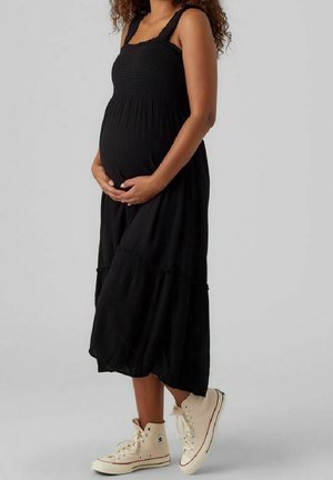 Mujer embarazada con vestido midi negro sin mangas y zapatillas beige de caña alta, de pie frente a un fondo gris claro y liso.