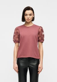 Γυναίκα που φοράει ροζ-σκουρόχρωμο τοπ με κοντά μανίκια και διάφανα μανίκια με κεντημένα λουλούδια, καθώς και μαύρο δερμάτινο παντελόνι, στέκεται μπροστά σε απλό φόντο.