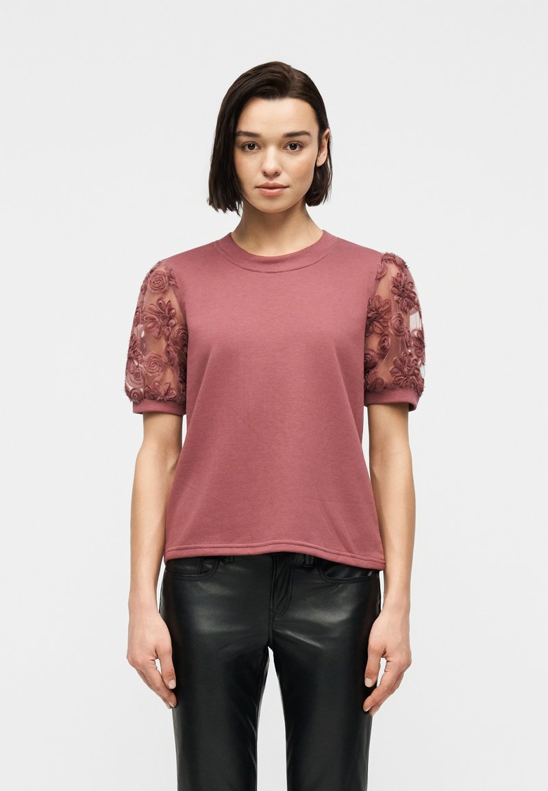 Γυναίκα που φοράει ροζ-σκουρόχρωμο τοπ με κοντά μανίκια και διάφανα μανίκια με κεντημένα λουλούδια, καθώς και μαύρο δερμάτινο παντελόνι, στέκεται μπροστά σε απλό φόντο.