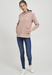 Sudadera rosa con un patrón texturizado, capucha con cordón y bolsillo en la parte delantera. Combinada con jeans skinny oscuros y zapatillas blancas.