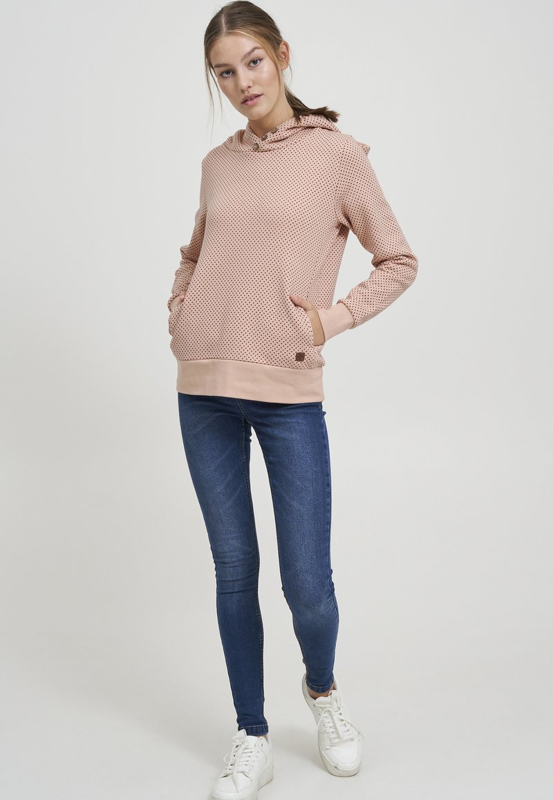 Sudadera rosa con un patrón texturizado, capucha con cordón y bolsillo en la parte delantera. Combinada con jeans skinny oscuros y zapatillas blancas.