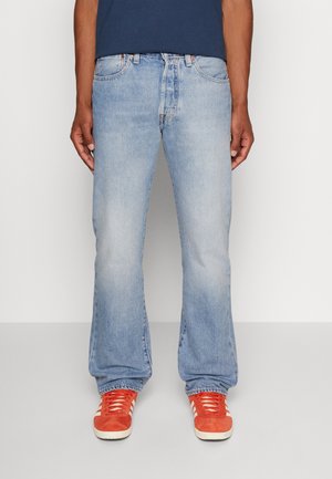 Homme portant un jean droit bleu clair et des baskets orange vif, debout devant un fond gris uni.