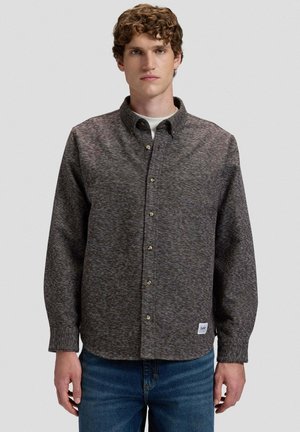 Junger Mann mit lockigem Haar, trägt ein graues, strukturiertes Hemd über einem weißen Unterhemd und blauen Jeans, steht vor einem schlichten Hintergrund.