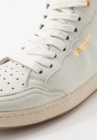 Cremefarvet læder sneakers med perforeret tå, flade hvide snørebånd, guldbelægning og struktureret beige gummisål. Rund form med syede detaljer.