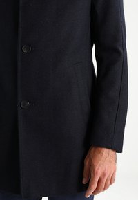 Primer plano de una persona con un abrigo de lana azul marino oscuro, con botones delanteros y un bolsillo lateral, con la mano relajada a su lado.