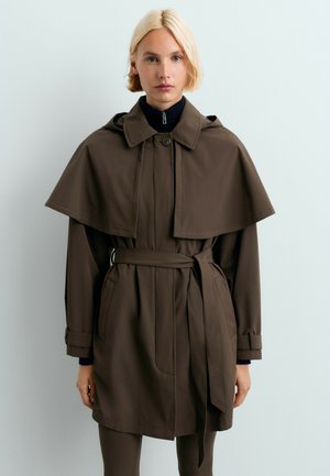 Femme blonde portant un trench-coat marron foncé ceinturé avec un détail cape et un haut noir à col montant en dessous.