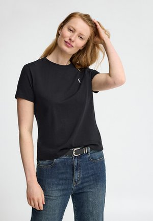 Kobieta z jasnoczerwonymi włosami nosi czarną koszulkę z krótkim rękawem i niebieskie dżinsy, dotykając włosów jedną ręką na jednolitym jasnym tle.