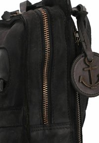 Bolso de cuero negro con una superficie texturizada, que presenta un cierre de cobre prominente y una etiqueta de cuero redonda con un emblema de ancla.