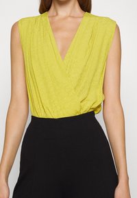 Haut jaune sans manches avec un profond décolleté en V, présentant un motif de logo subtil et des détails froncés à la taille, associé à un pantalon noir taille haute.