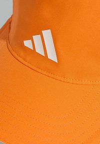 Oranžový klobouk s hladkou texturou. Má tři bílé pruhy s logem. Klobouk má prošitou krempu a je vyroben z měkké, odolné látky.