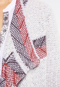 Pletený kardigan se světle šedým základem, s červeným a tmavě šedým vzorovaným akcentem na límci, texturovanou látkou a stříbrnými knoflíky.