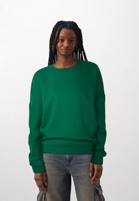 Sudadera verde de gran tamaño en una tela suave, con escote redondo y puños acanalados. Combinada con pantalones de mezclilla claros que tienen bolsillos tipo cargo.