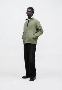 Olive green jacket with a zip pocket, collar design, and black pants. Smooth texture, lightweight material, paired with black shoes.