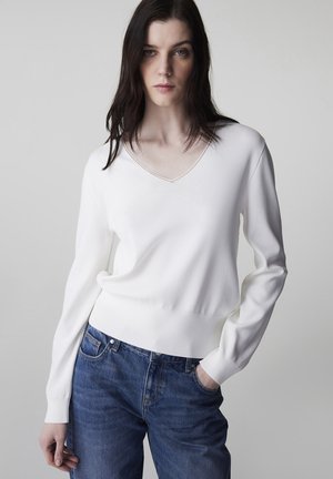 Junge Frau mit langen dunklen Haaren, die einen weißen V-Ausschnitt-Pullover und blaue Jeans trägt, posiert mit einer Hand in der Tasche vor grauem Hintergrund.