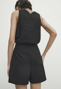 Haut noir sans manches avec un dos en V, associé à un short noir sur mesure doté d'une poche latérale et d'une texture de tissu lisse.