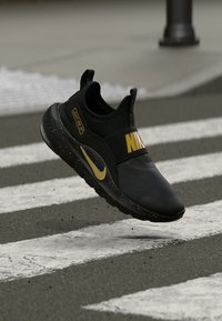 Schwarzer Nike-Sneaker mit goldenem Logo und Akzenten, ausgestattet mit einem Mesh-Obermaterial, gepolstertem Kragen und gesprenkelter Sohle, platziert über einem Zebrastreifen.