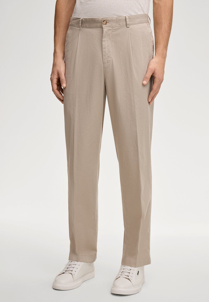 Pantalon beige à coupe décontractée, avec des plis, une fermeture à bouton et des poches latérales. Texture de tissu doux associée à des baskets blanches.
