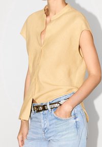 Camicia a maniche corte in lino giallo chiaro, con scollatura a V e chiusura a bottoni, abbinata a jeans in denim azzurri e una cintura borchiata.