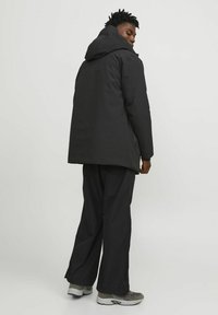 Chaqueta negra con capucha y acabado mate, de mangas largas y corte relajado, combinada con pantalones negros holgados y zapatillas grises.