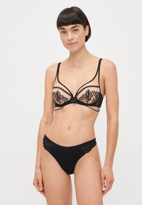 Μαύρο σετ εσωρούχων που περιλαμβάνει ένα διαφανές bralette με λουλουδάτη κεντητική, σκοπίδια και αντίστοιχες χαμηλοκάβαλες σλιπ με υφασμάτινες λεπτομέρειες.