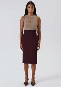 Donna in piedi che indossa un top senza maniche beige a maglia con ritagli, una gonna a matita alta di vita color viola scuro con bottoni e tacchi a punta.
