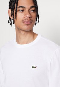 T-shirt à manches longues blanche avec un petit logo de crocodile brodé en vert sur la poitrine. Matière en coton doux avec un col rond classique.