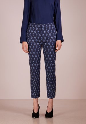 Mujer con top de manga larga azul marino y pantalones hasta el tobillo con patrón azul, con zapatos de tacón alto negros, de pie sobre un fondo liso.