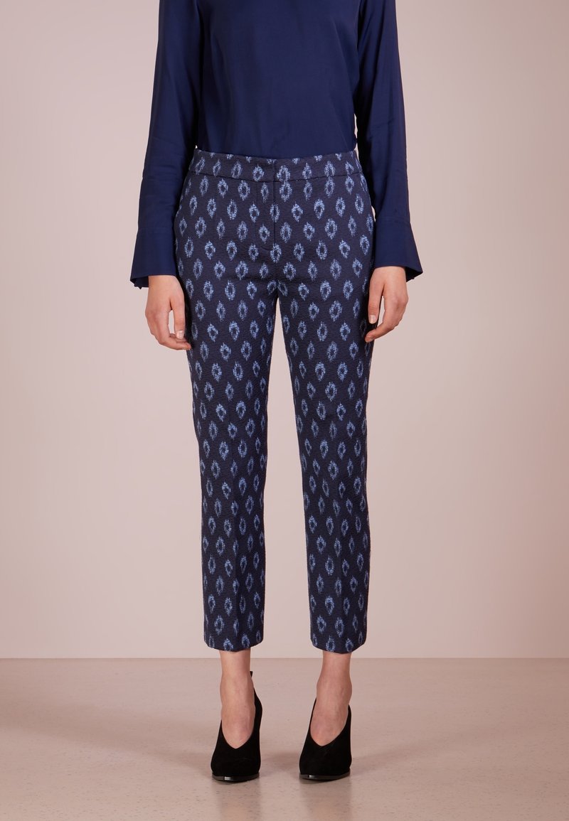 Femme portant un haut bleu marine à manches longues et un pantalon bleu à motifs longueur cheville avec des chaussures à talons noirs, debout devant un fond uni.