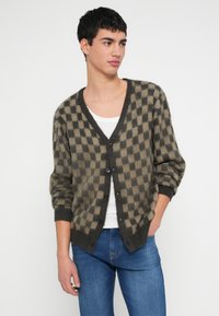 Młoda osoba ubrana w kraciasty brązowo-beżowy sweter zapinany na guziki, pod którym ma biały top na ramiączkach i niebieskie dżinsy, stoi z jedną ręką w kieszeni.