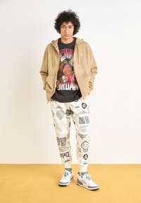 Sudadera con cremallera de color marrón claro sobre una camiseta negra con gráfico, combinada con pantalones blancos que tienen logotipos y detalles en negro, y zapatillas gris y blancas.