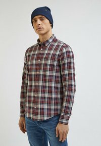 Koszula w kratę w kolorze czerwonym, granatowym i białym z kołnierzykiem zapinanym na guziki i jedną kieszonką na piersi. Nosi ją w połączeniu z granatową dzianinową czapką. Widoczne niebieskie dżinsy.