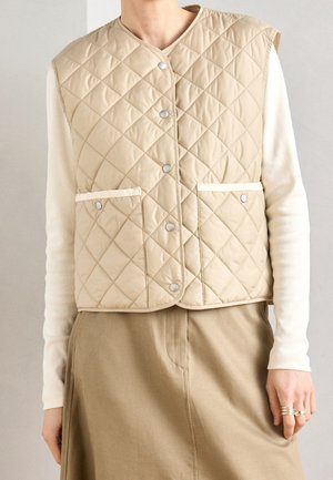 Person iført en beige quiltet vest med trykknapper og forlommer over en langærmet cremefarvet top og matchende beige nederdel.