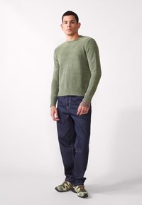 Green knitted sweater with a crew neckline, paired with dark blue denim jeans. Model wears patterned green and black athletic shoes.