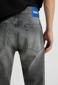 Gray denim jeans with a faded finish. Features a blue logo patch on the waistband and two back pockets with stitched edges.