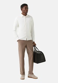 Weißes Hemd mit Knöpfen, beige Hosen, beige Sneaker, hält eine schwarze Tasche. Klare, moderne Gestaltung mit glatten Texturen und neutralen Farben.