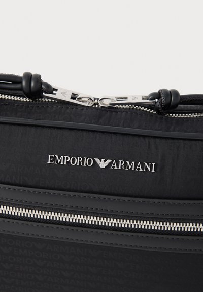 Juodos spalvos nailoninė rankinė su Emporio Armani logotipu, sidabrinėmis detalėmis ir užtrauktuku, kuriame matomas kontrastingas siuvimas. Matyti tekstūruotas paviršiaus raštas.