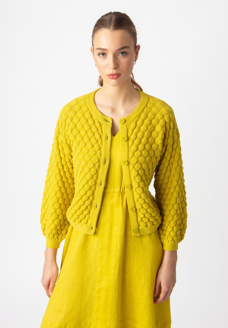 Kollane kootud kardigan tekstuursete pallikesedetailidega, ribakujulised varrukad ja viie nupuga. Kombineeritud sobiva kollase kleidiga.