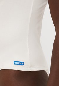 Balti juostelėmis marškinėliai su prigludusia danga, šone turi mėlyną Adidas logotipo etiketę. Lygus paviršius, be matomų raštų.