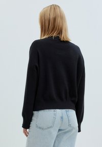 Schwarzer Strickpullover mit bequemer Passform, fallenden Schultern und geripptem Saum. Kombiniert mit hellblauen, hochgeschnittenen Jeans.