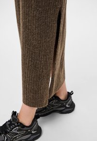 Jambe inférieure portant une jupe côtelée marron avec une fente sur le côté, associée à des baskets de sport noires et grises sur fond blanc.