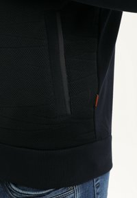 Primer plano de una persona que lleva una chaqueta oscura texturizada con ribete acanalado y bolsillo lateral con cremallera, combinada con jeans de mezclilla azules.