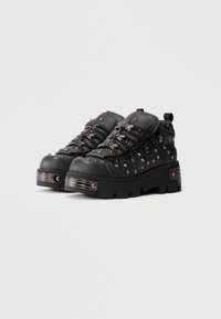 Sapatos altos texturizados pretos com adornos de tachas prateadas e ferragens decorativas em metal. Apresentam uma sola grossa e atacadores contrastantes.