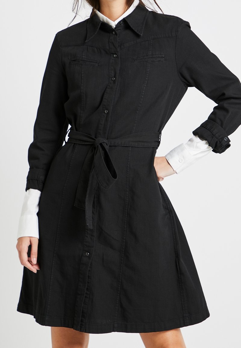 Femme portant une robe noire boutonnée avec une ceinture à la taille et des poignets de chemise blancs visibles sous les manches longues.