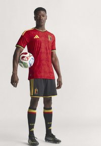 Crvena nogometna majica s teksturiranim uzorkom, crne kratke hlače i crne čarape s crvenim, žutim i crnim prugama. Brendiranje Adidas prisutno.