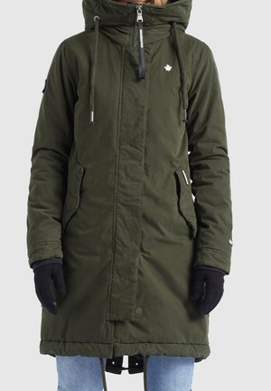 Langer olivgrüner Wintermantel mit Kapuze, Reißverschluss- und Knopfverschluss, Seitentaschen, schwarze Handschuhe und hellblaue Jeans an einer Person.