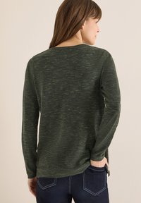 Langærmet grøn sweater med tekstureret mønster. Rund halsudskæring og sideslids ved kanten. Båret med mørke jeans, set bagfra.